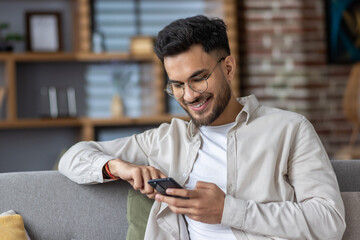 Smiling man using smartphone while relaxing on couch at home, enjoying digital content and staying...