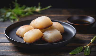A plate of steamed buns sits on a wooden table with a bowl of dipping sauce in the background