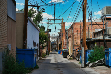 An urban alley adorned with numerous power lines stretches into the distance, flanked by a variety of blue dumpsters and aged brick buildings under a bright, clear sky.