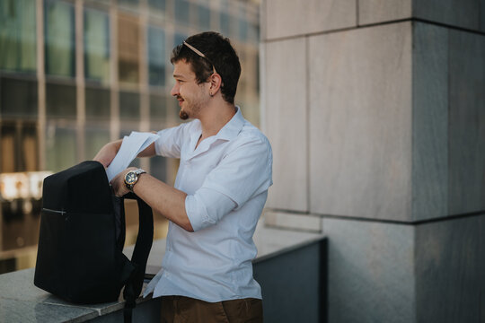 Professional young man in casual attire organizing documents in his backpack outside a modern office building. Represents business, organization, and efficiency in an urban environment. - Powered by Adobe