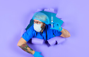 Doctor with stethoscope in medical mask and hairnet with syringe looking through paper hole. Male...