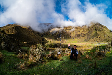 Hispanic traveler waving while crouching in a mountainous valley, surrounded by green hills and low clouds, with a waterfall and a partially clear sky in the background.