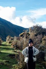 Portrait of uncomfortable latin woman with arms on waist wearing black beanie and beige jacket against green hills background