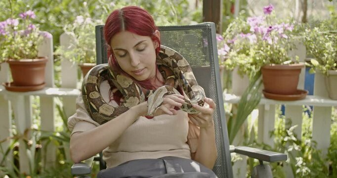 A woman sits in a garden with her pet snake around her shoulders.