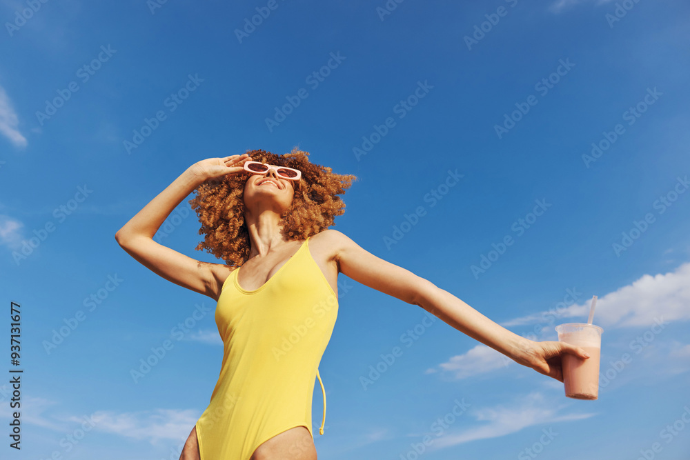 Wall mural a joyful woman in a yellow swimsuit enjoying a sunny day at the beach with a drink in hand, smiling 