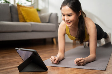 Happy Asian slim woman do routine exercise training plank at home apartment doing stretching yoga posture and watching video on smart pad with online lesson course personal trainer in the living room