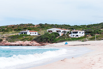 Fototapeta premium Overlooking the bay from Isola Rossa beach, the turquoise waters gently lap against the pink rocky coastline, where a few Mediterranean villas are nestled among the lush green hills. 