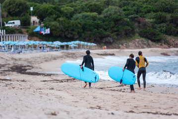 A trio of young surfers, clad in wetsuits and carrying their boards, walk together after emerging...
