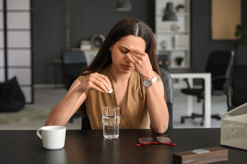 Young woman with glass of water and soluble tablet suffering from headache sitting in office