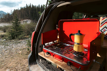 view of a metal camping stove in the back of a van with a yellow coffee maker in the sun light