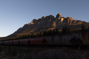 view of a long cargo train passing by in a national park with tall mountains behind in summer with...