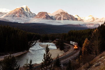 view of a long train with a red locomotive with the lights on following a curved river on a sunrise with low clouds over the forest and rays of lights on top of the mountains