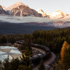 view of a long train with black wagons following a curved river on a sunrise with low clouds over...