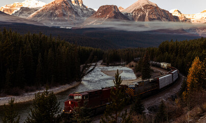 view of a long train with wagons and a red locomotive following a curved river on a sunrise with...