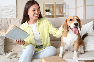 Young woman with cute Beagle dog reading book on sofa in living room