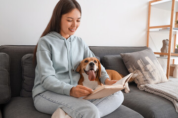 Young woman reading book and sitting with adorable Beagle dog on sofa in living room