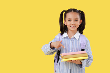 Cute little happy Asian girl with backpack pointing at books on yellow background. End of school...