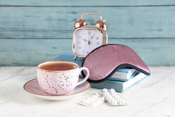 Cup of tea with sleep mask, books, pills and alarm clock on white table