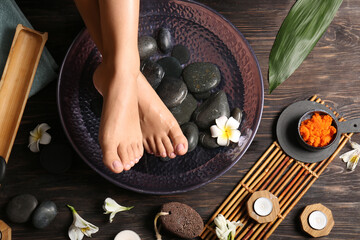 Female legs in bowl for spa procedure with water, flowers and stones on wooden background