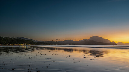 Twilight after sunset on a tropical island. The blue sky is highlighted with orange at the horizon. Silhouettes of mountains. Ocean waves are spreading over the beach. Reflection on wet sand. Lights
