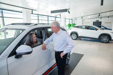 An elderly Caucasian woman is sitting in a new car. Her husband is standing nearby.