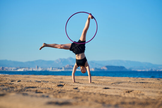 A person performs a handstand with a hula hoop on a beach