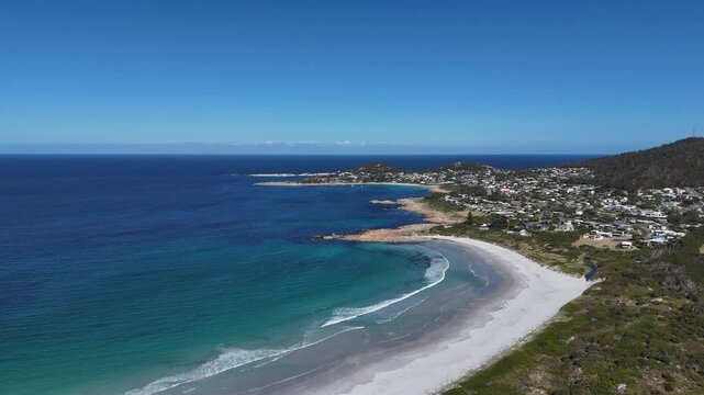 Aerial flight along sandy beach of Tasmania with Bicheno Town in background. Blue ocean and sky during sunny day. Wide shot.