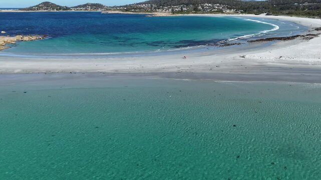 Aerial flyover turquoise water of ocean in Tasmania. Walking people on sandy beach connecting Diamond Island. Bicheno City in background. Holiday trip in summer.