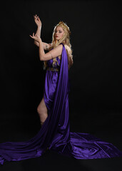 full length portrait of beautiful blonde female model wearing purple and gold silken toga robes with royal crown, historical fantasy goddess costume. Isolated on dark studio background.