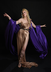 full length portrait of beautiful blonde female model wearing purple and gold silken toga robes with royal crown, historical fantasy goddess costume. Isolated on dark studio background.