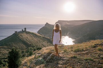 Fototapeta premium A blonde woman stands on a hill overlooking the ocean. She is wearing a white dress and she is enjoying the view.