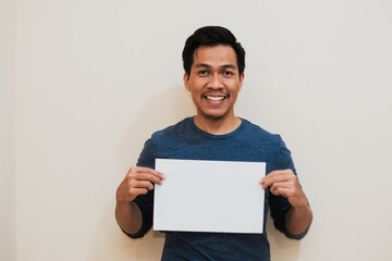 Happy Southeast a Asian man wearing long blue sleeve holding a piece of blank sheet of paper looking at the camera and smiling on cream background. Job search, sale concept