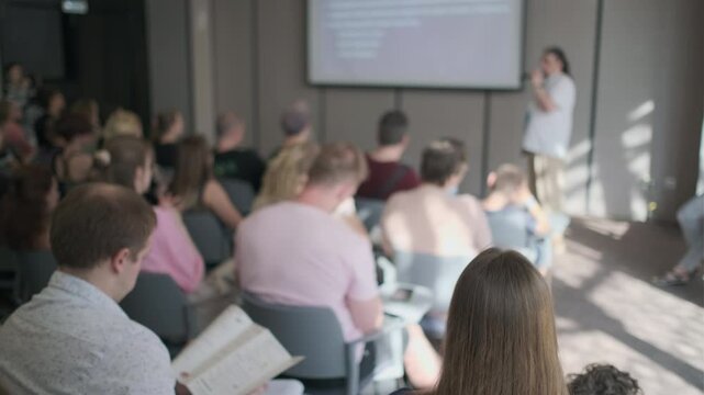 Diverse group of people attentively listening to a presentation in a well-lit conference room. The atmosphere is professional and focused, with attendees taking notes.