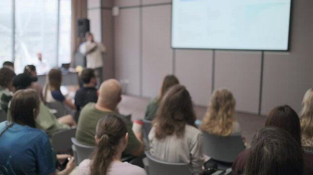 Group of people attentively listens to a presentation in a modern conference room. The speaker is presenting at the front with a large screen displaying visuals.
