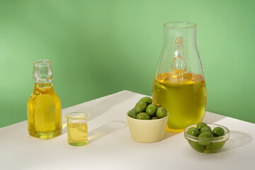 Minimal concept with bowl and glass flasks filled with olives and olive oil on the green background. Scene for advertising. The bark, leaves and oil of olive peel all have many benefits