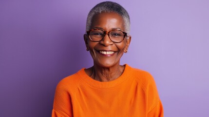 A cheerful senior black woman beams with joy while wearing stylish glasses and an orange sweater. The vibrant purple backdrop enhances her warm smile