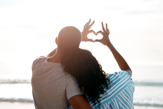 Hands, heart shape and back with couple, beach and happy people for anniversary on holiday together. Romance, African man and woman with ocean, summer break and relax with sunset embrace on vacation
