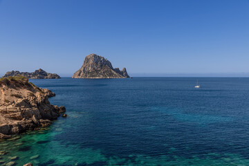 Es Vedra island in Ibiza, Spain, with a catamaran sailing on the clear blue waters. The rocky...