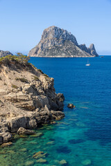 Es Vedra off Ibiza, Spain, rises dramatically from the Mediterranean Sea. A sailboat is anchored in the vibrant blue waters near the rocky coastline