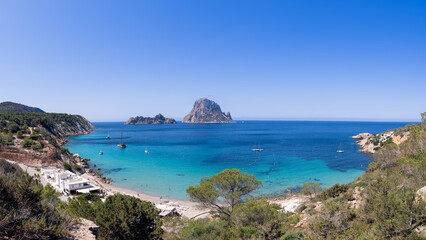 Expansive panorama of Cala d'Hort beach, Ibiza, showcasing Es Vedra island, tranquil turquoise waters, sailboats, and surrounding cliffs with vegetation, all beneath a bright sky