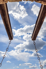 Supporting beams of a drawbridge in a medieval castle in Brescia, Italy, featured in a vertical conceptual photo. The beams and chains are set against a backdrop of blue sky and clouds