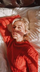 Fototapeta premium A stylish blonde woman relaxes on a bed, wearing a cozy red sweater. Sunlight casts delicate shadows across her face, enhancing the calm morning ambiance