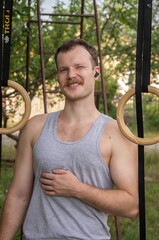Obraz premium Winking man with a mustache standing between wooden gymnastic rings, wearing a gray tank top and wireless earbuds in an outdoor setting with trees.