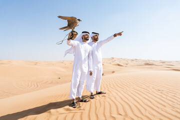 Two middle-eastern emirati men wearing arab kandura holding falcon in the desert