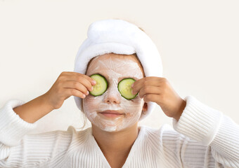 A little girl with a white beauty mask on her face holds fresh cucumbers in front of her eyes. On a white background. Beauty Treatment Spa Concept
