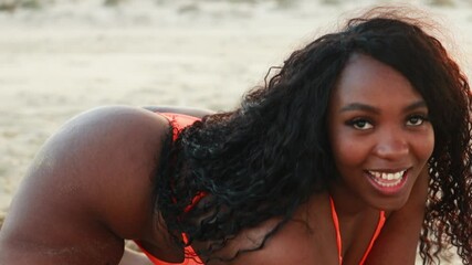 Happy overweight woman sitting on beach in swimsuit on sand dancing. Summer vacation concept of dark-skinned girl, body positivity