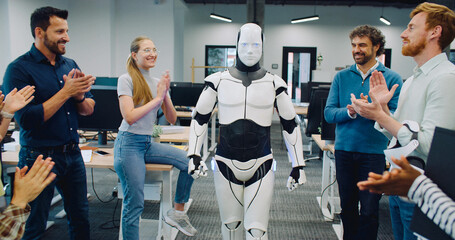Robot walking in middle of room. Group of multi-ethnic coworkers clapping.