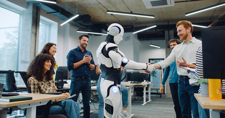 Multi-ethnic group of people surrounding their new creation in middle of large office room. Clapping together. In middle robot dancing before giving handshake to male. Showing thumbs up to camera.