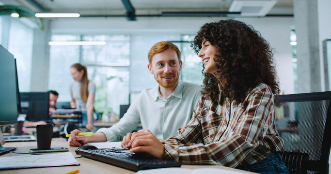 Camera moving closer to two Caucasian colleagues working side by side in modern office. Woman typing on keyboard. Man showing something on notepad. Looking at monitor. Controlling new intern.