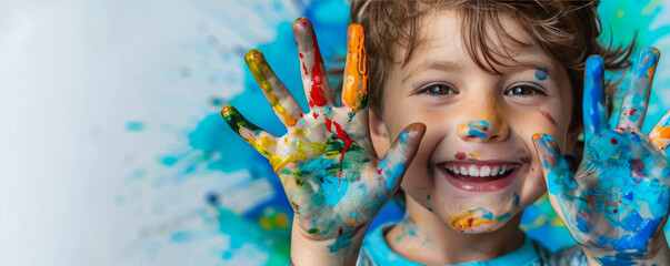 A joyful child with colorful painted hands smiles brightly against a vibrant blue background, showing creativity and fun during an arts and crafts activity. banner with place for text